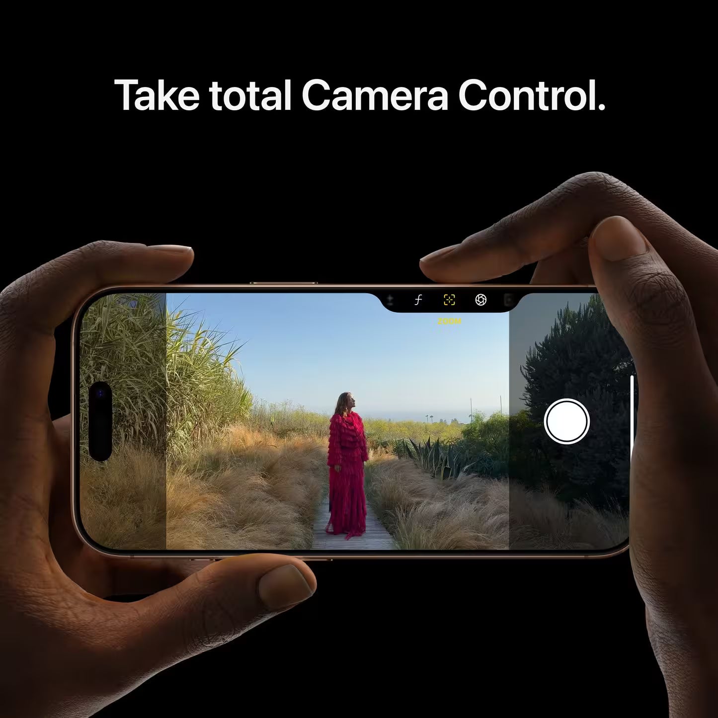 A person holding the iPhone 16 Pro Max in Desert Titanium in landscape mode, with their index finger using the Camera Control button, showing the camera interface and a woman in a red dress, set against a black background.